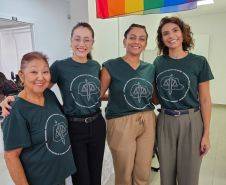 Imagem de uma foto espontânea de servidoras públicas, todas usando uma camiseta verde da Defensória Pública do Estado do Paraná. No fundo da imagem, dá para ver uma bandeira LGBTQIA+ pendura do teto.