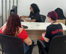 Imagem que mostra duas pessoas sendo atendidas no mutirão em Ponta Grossa. Elas estão de costas e entre elas, é possível ver a pessoa que está fazendo o atendimento, sentada atrás da mesa e olhando para a tela do computador.