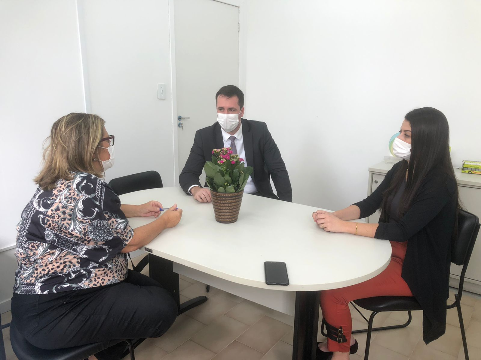 A foto mostra três pessoas, um homem e duas mulheres, sentados à mesa, usando máscaras