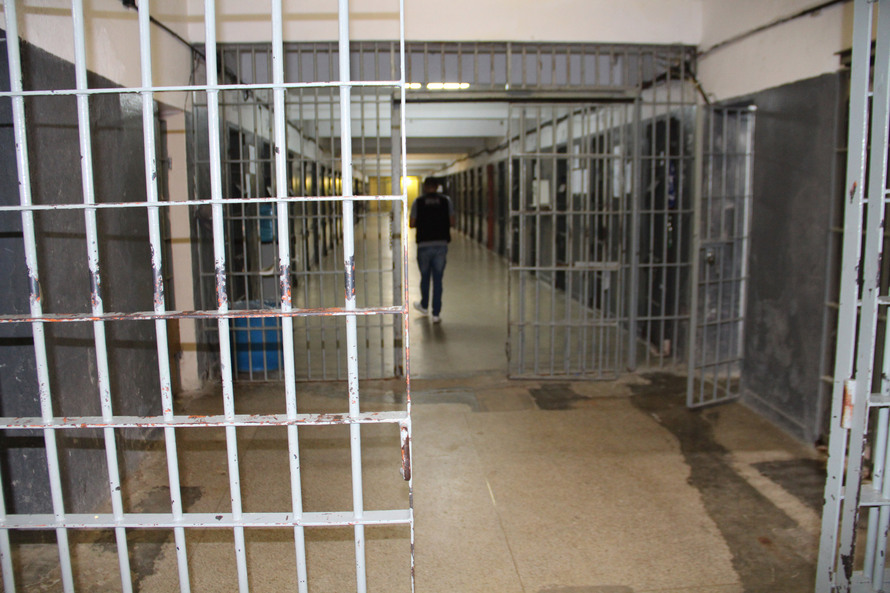 Foto mostra o interior de uma prisão, com um agente penitenciário de costas e um cela com a porta aberta