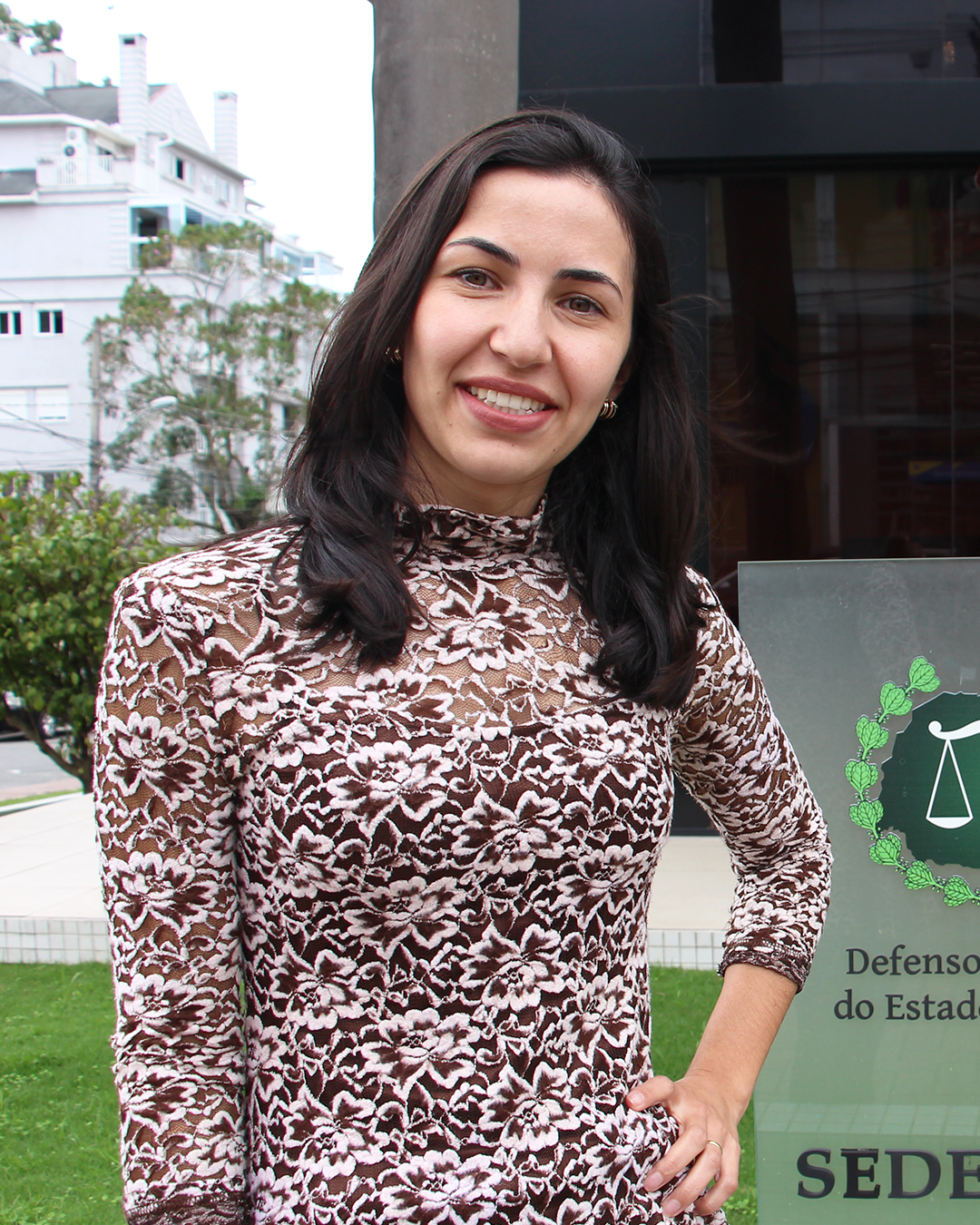 Imagem que mostra a servidora Jeniffer dos Santos em frente à sede administrativa da Defensoria Pública. Ela sorri para câmera, está com os cabelos soltos e uma blusa de mangas longas, de renda, em tons terrosos. Ela está com uma das mãos na cintura.