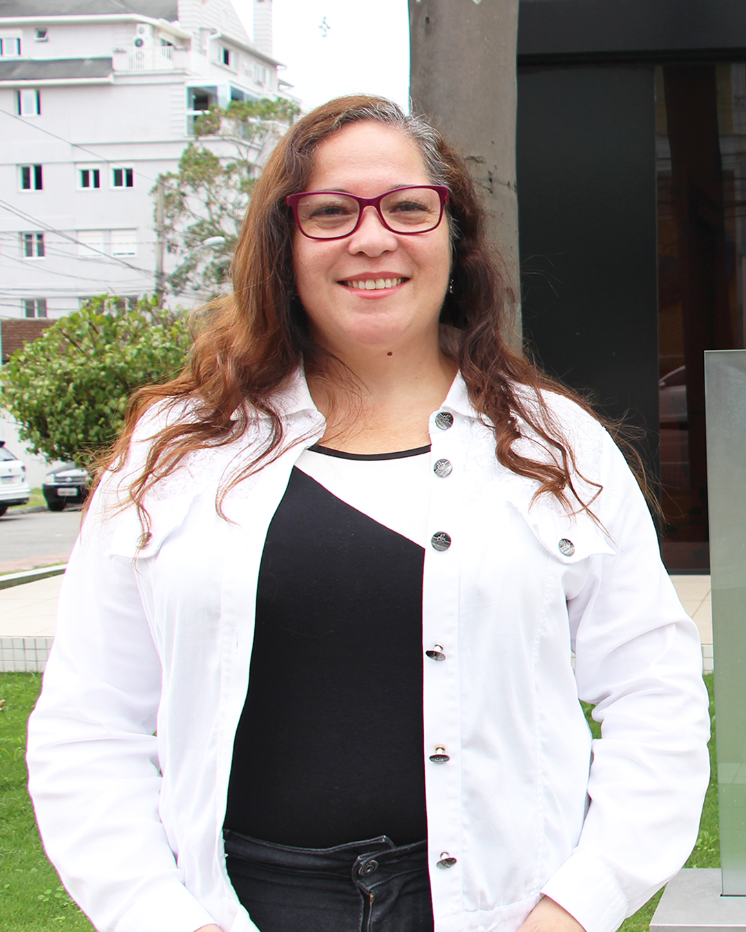 Imagem que mostra a servidora Rosemeri Aparecida e Silva em frente à sede administrativa da Defensoria. Ela usa uma jaqueta branca sobre uma blusa preta com detalhe branco. Está de óculos, cabelos soltos e sorri para a foto.