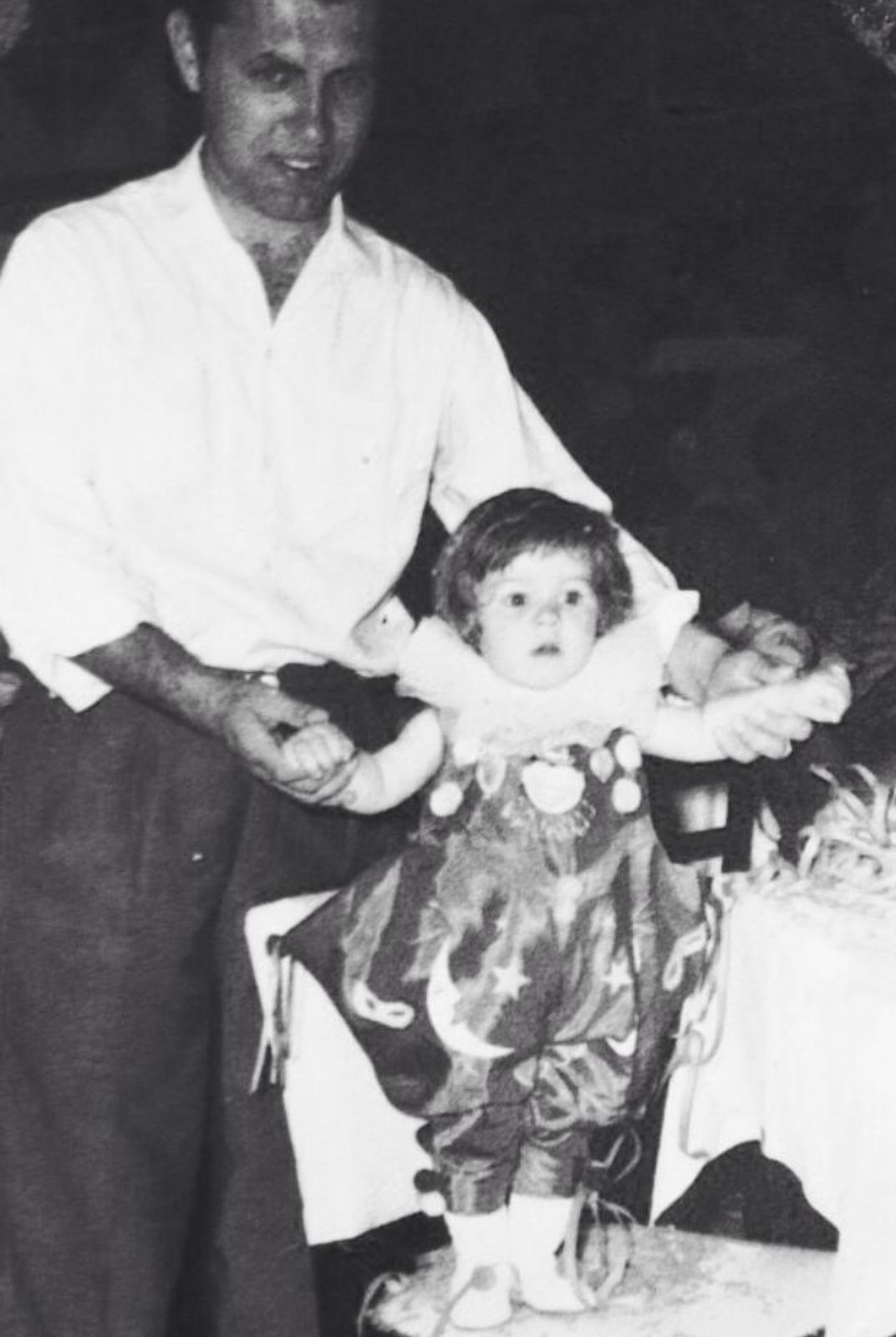 Imagem preta e branca do pai de Denise segurando ela quando era bebê. Ele segura em suas mãos e ela está em pé em cima de uma mesa.