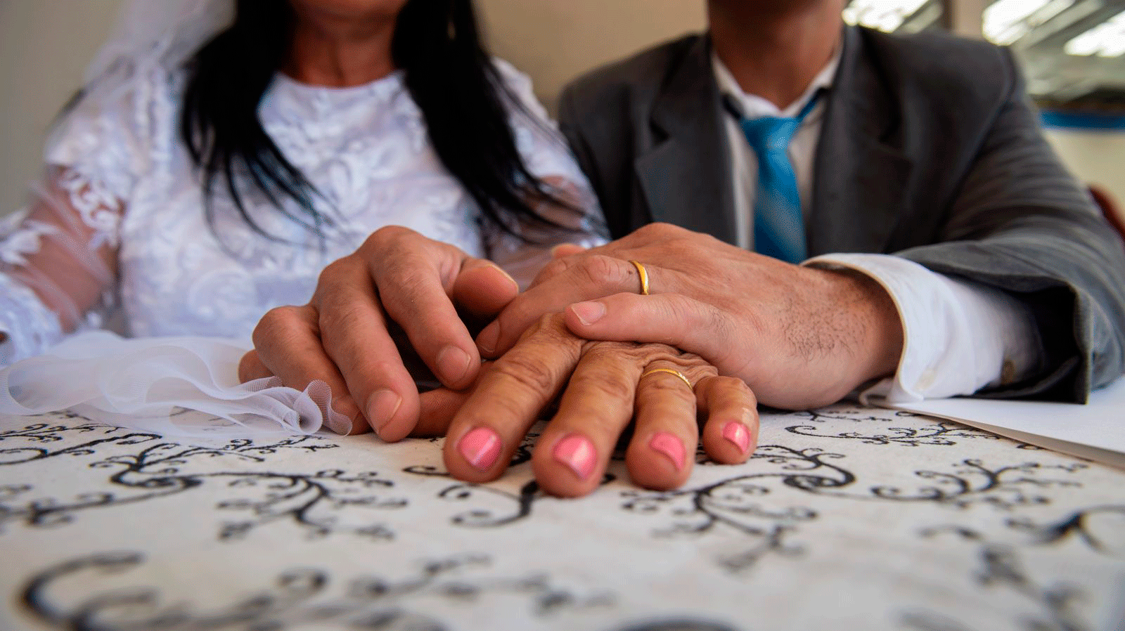 casal com as mãos sobre a mesa mostrando as alianças. eles usam trajes de terno e gravata e vestido de noiva.