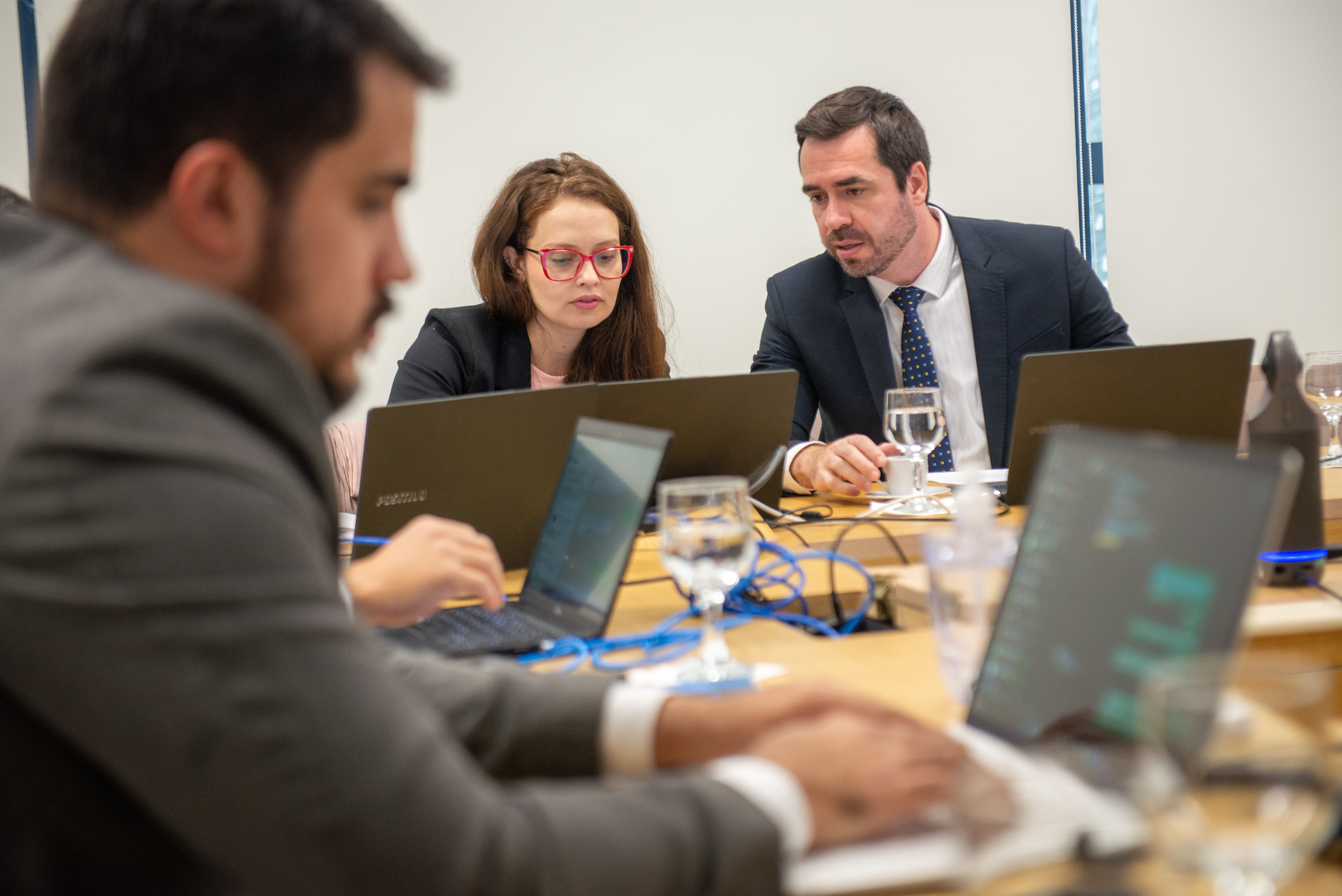 Imagem de um homem de terno em primeiro plano e ao fundo um homem de terno e uma mulher de óculos vermelho conversam olhado a tela de um laptop.