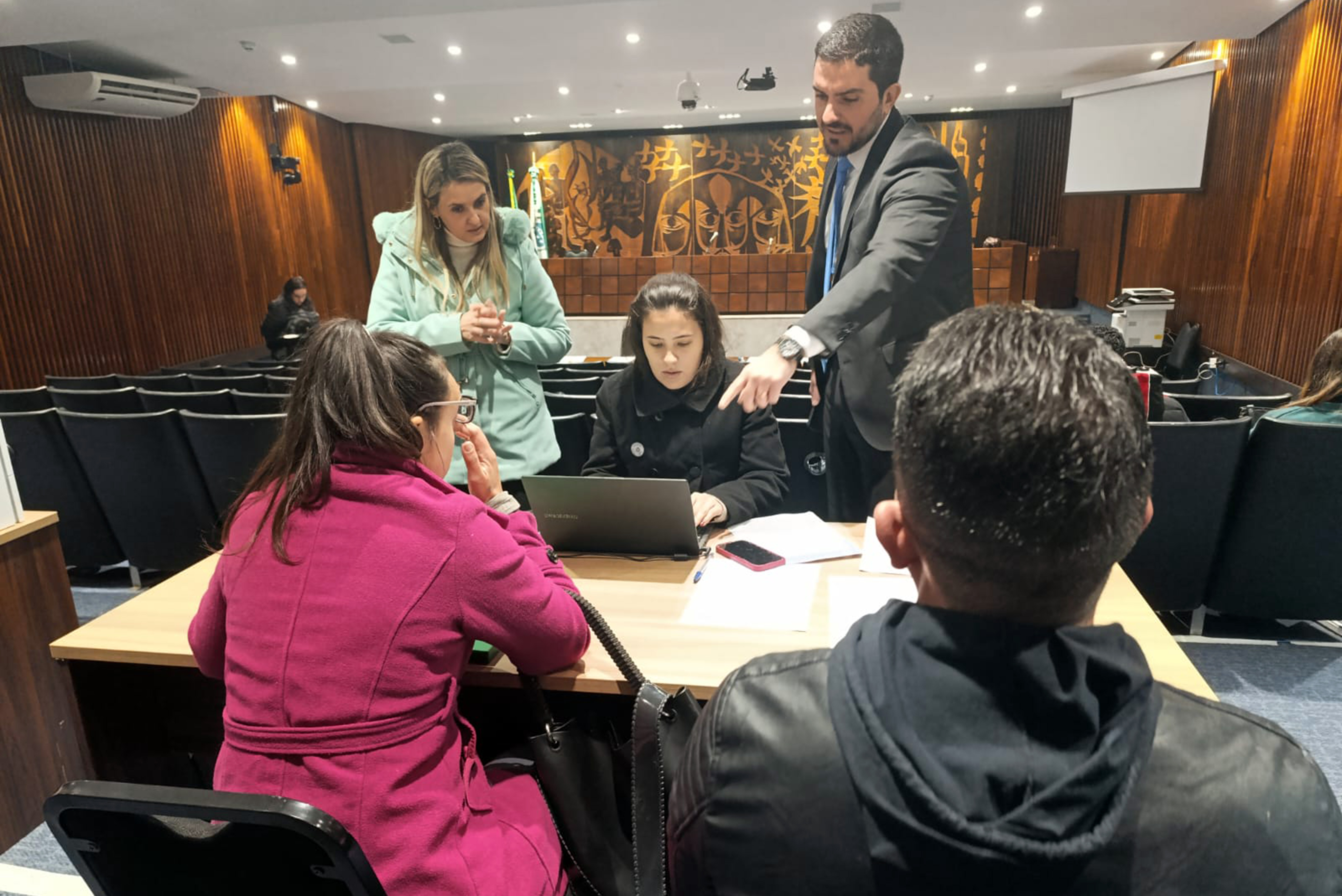 De costas um casal sentado recebe orientações da equipe da DPE-PR, formada por duas mulheres e um homem. Ele aponta a tela de um computador.