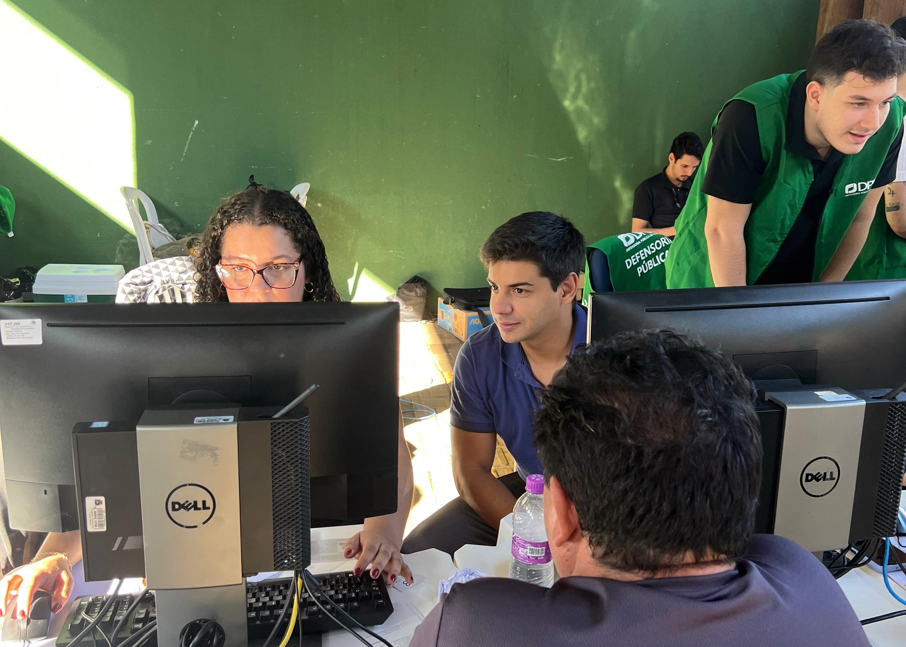 Imagem mostra três servidores utilizando computadores durante os atendimentos do mutirão. De costas em primeiro plano se vê um assistido.