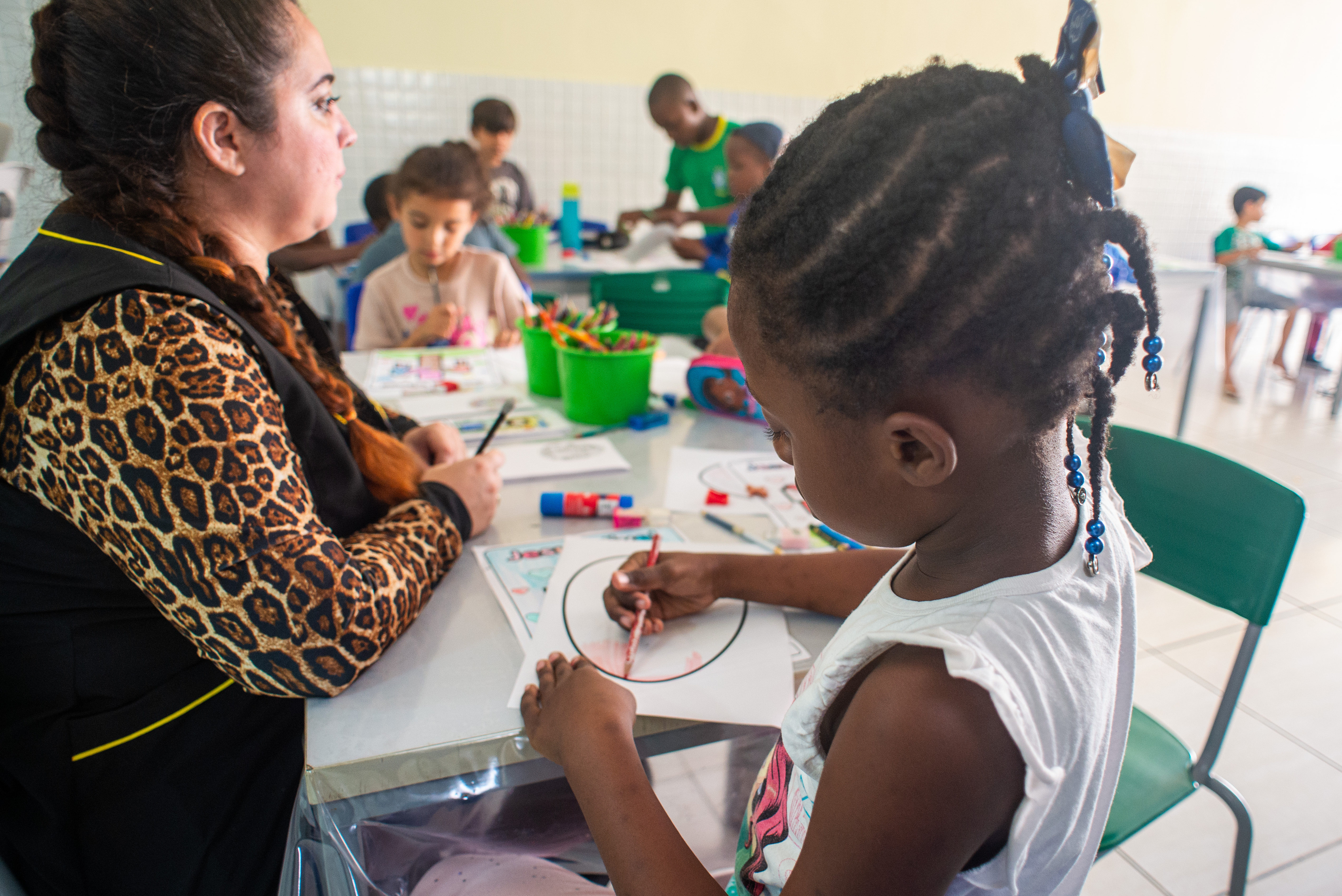 Imagem com crianças sentadas, junto de uma professora, enquanto realizam atividades escolares. O rosto das crianças está borrado e a criança em evidência está de costas.