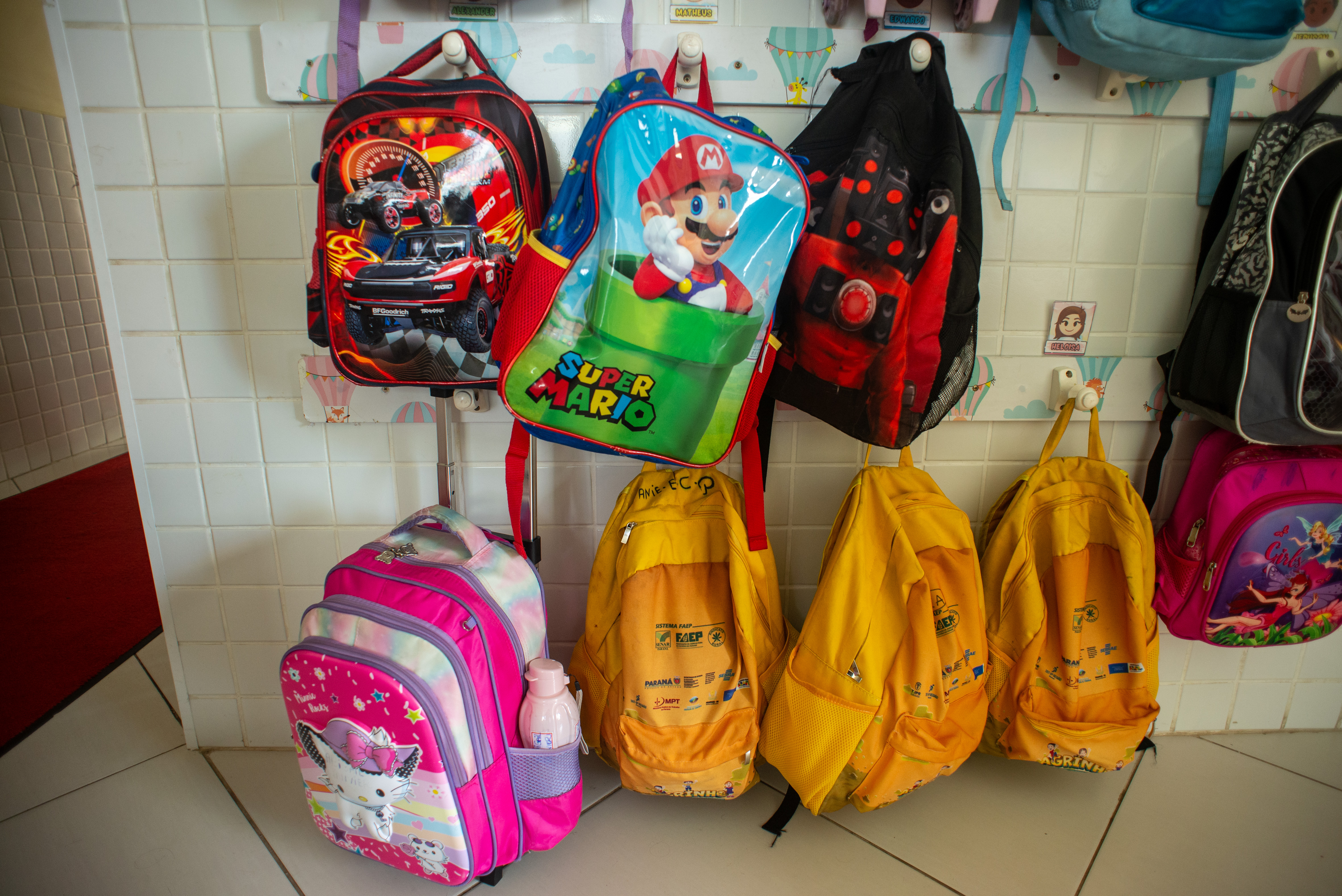 Imagem com várias mochilas de crianças em um local de estudos. Elas estão todas penduradas em ganchos na parede.