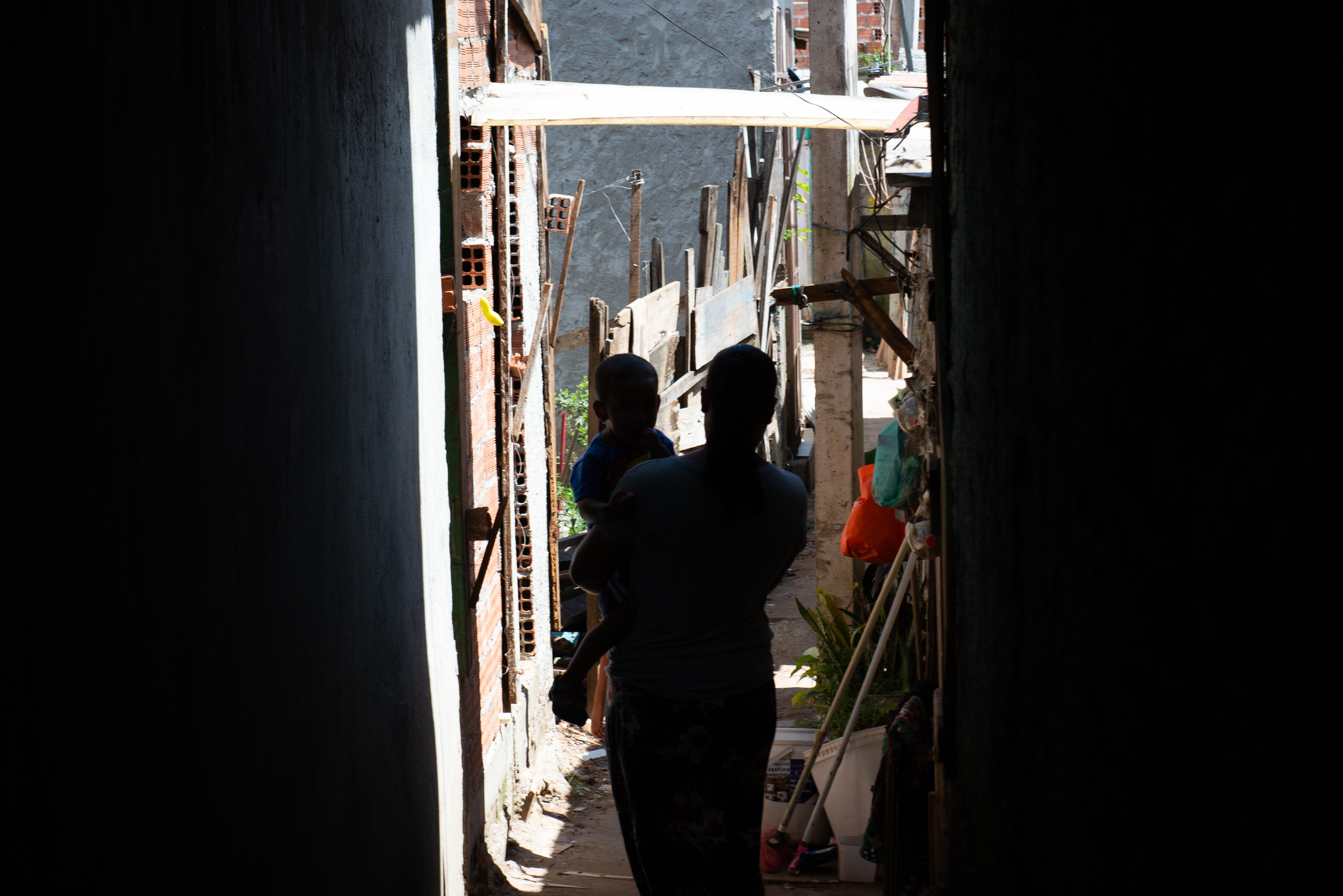 Imagem com uma mulher segurando uma criança no colo. Elas estão em silhueta de sombra, em um corredor com a luz apenas ao final da imagem.