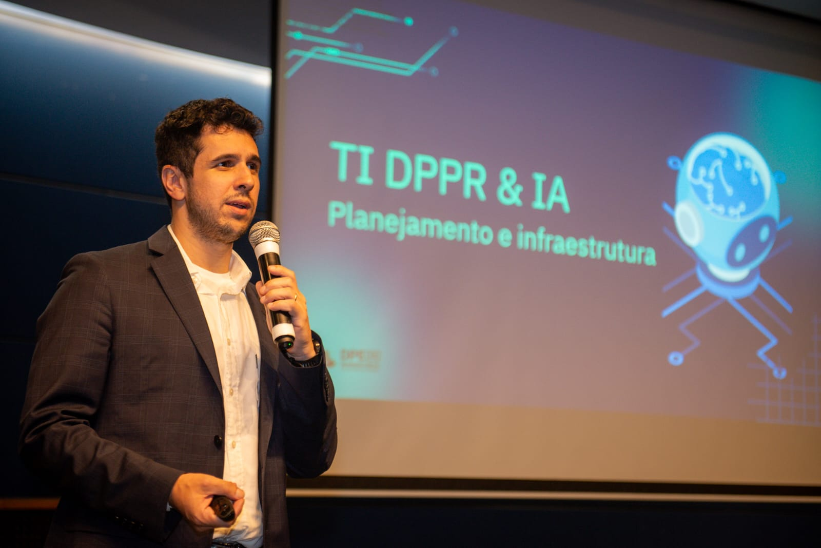 Imagem do defensoria Nicholas Silva apresentando o projeto de Interligencia Artifical na Defensoria em um evento sobre tecnologia. Na foto, ele está em um palco, ao lado de uma tela de projeção segurando um microfone.