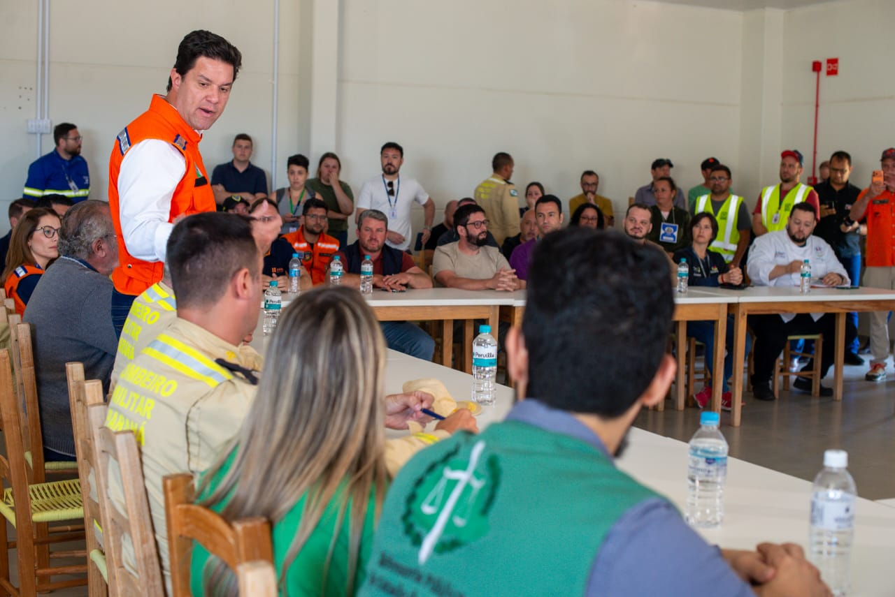Em uma sala de reunião, o secretário das cidades do Estado, Guto Silva, está em pé vestindo colete laranja (Defesa Civil/Casa Militar), fala com o público sentado nas mesas brancas. Em primeiro plano, uma mulher e um homem de colete verde da Defensoria Pública ouvem.