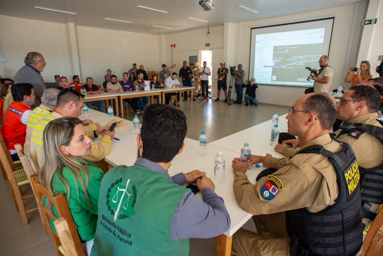 Cena de reunião com várias pessoas sentadas em mesas brancas em "U" em uma sala iluminada. Em primeiro plano, defensores públicos juntos de oficiais da Polícia Militar e outras autoridades, sentados. Há uma tela de projeção exibindo um mapa ao fundo.