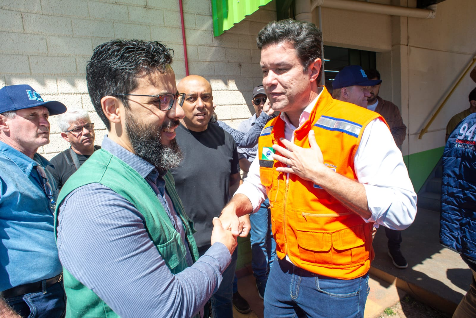 Dois homens brancos se cumprimentam com um aperto de mãos. O da esquerda, o defensor público Fernando Redede, com barba e óculos, veste um colete verde da defensoria. À direita, o secretário das cidades do Estado, Guto Silva, veste um colete de segurança laranja, com a mão esquerda sobre o peito. Há outras pessoas ao fundo.
