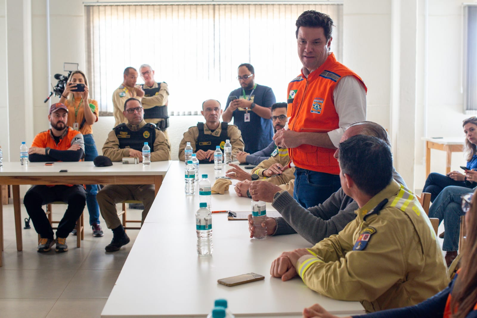 Em uma sala de reunião, o secretário das cidades do Estado, Guto Silva, está em pé vestindo colete laranja (Defesa Civil/Casa Militar), fala com o público sentado nas mesas brancas.