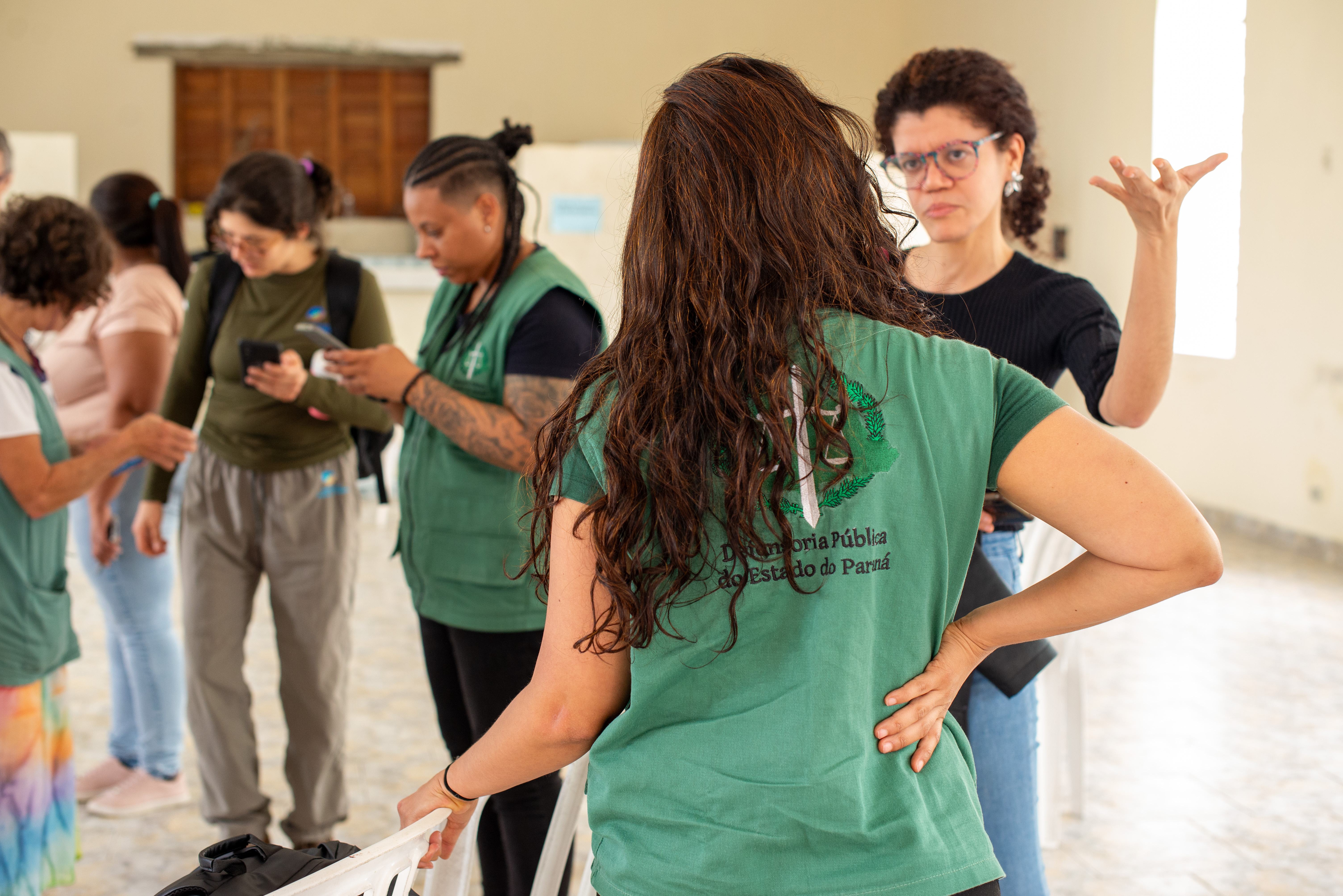 Equipe da Defensoria realiza atendimentos em localidade no Litoral do Paraná. Imagem mostra Defensora Pública de costas usando colete da instituição conversando com uma mulher. Ao fundo é possível ver mais servidores falando com pessoas em atendimento. 