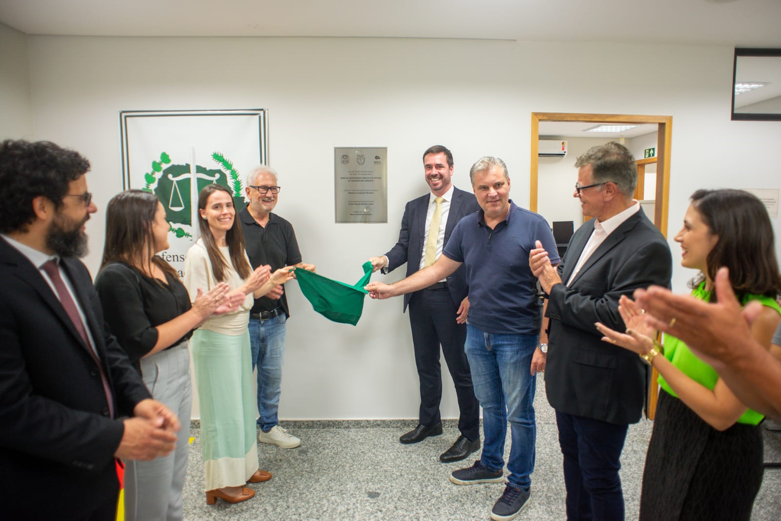 Um grupo de sete pessoas, entre homens e mulheres, sorri e bate palmas enquanto duas delas puxam um tecido verde para inaugurar uma placa prateada fixada na parede branca de um corredor.