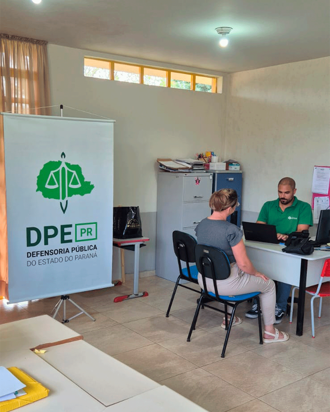 Visão mais ampla da sala com um banner vertical da DPE-PR em destaque à esquerda. Ao fundo, o funcionário de verde usa o notebook enquanto atende uma mulher de blusa cinza sentada à sua frente.