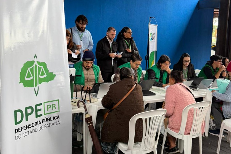 Em um espaço com parede azul, profissionais com coletes verdes usam notebooks em mesas brancas para atender cidadãos sentados à sua frente. À esquerda, destaca-se um banner vertical da Defensoria Pública do Paraná.