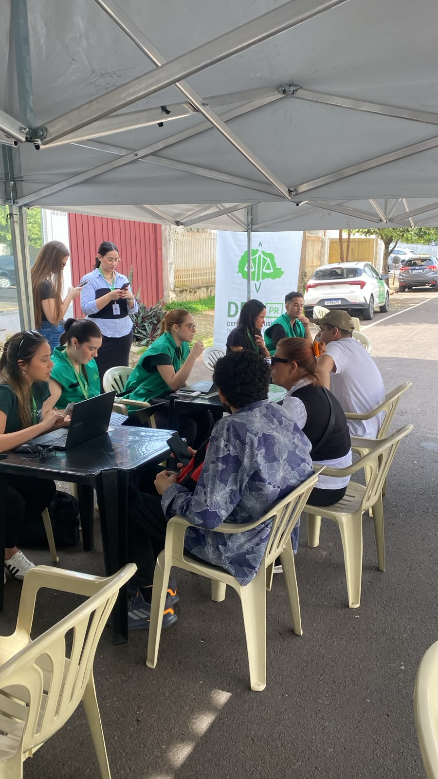 Em uma tenda de atendimento na rua a equipe da Defensoria Pública conversa e orienta as pessoas sentadas à frente das mesas. Ao fundo, destaca-se um banner branco e verde da DPE-PR.