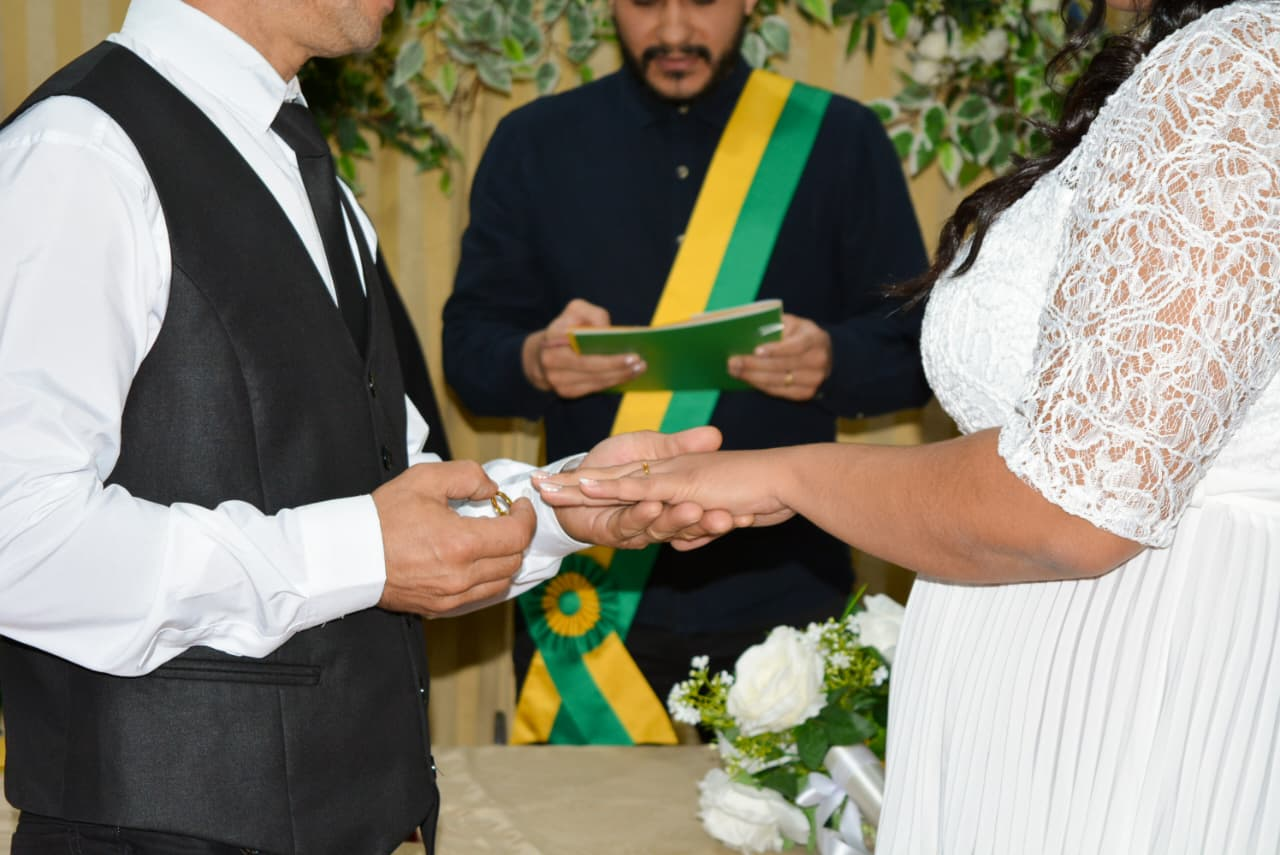 Detalhe do momento exato da troca de alianças. O noivo, de colete preto e camisa branca, coloca a aliança no dedo da noiva de vestido rendado, enquanto o celebrante com faixa verde e amarela observa desfocado ao fundo.