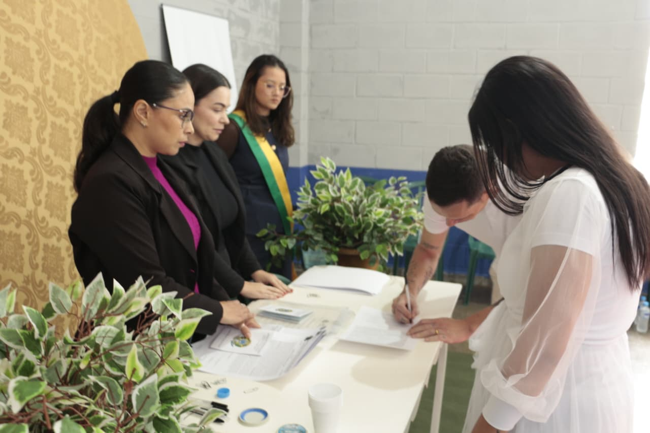 Um homem de camiseta branca assina um documento sobre uma mesa, ao lado de uma mulher de vestido branco. Atrás da mesa, três mulheres observam atentamente, sendo que uma delas usa uma faixa transversal com as cores da bandeira do Brasil.