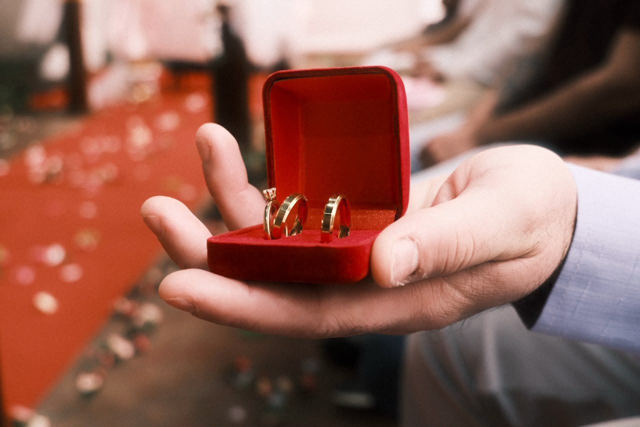 A mão de um homem, segura uma caixinha vermelha com três alianças de casamento. 