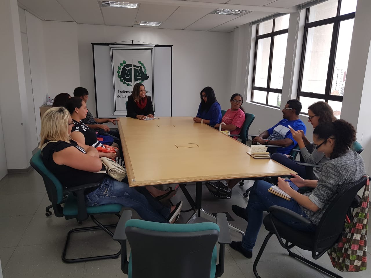 Mesa grande com representantes da Sociedade Barracão em volta e a dra. Olenka Lins ao fundo, do lado da bandeira da Defensoria Pública do Estado do Paraná.