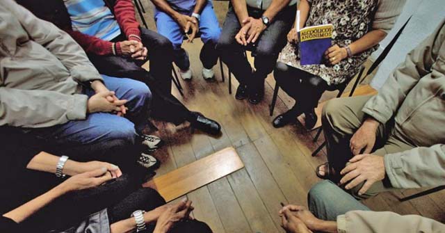 A foto foi tirada sob uma perspectiva de cima; Vemos um grupo de pessoas sentadas em roda, todas muito juntas, simulando uma reunião de AA.