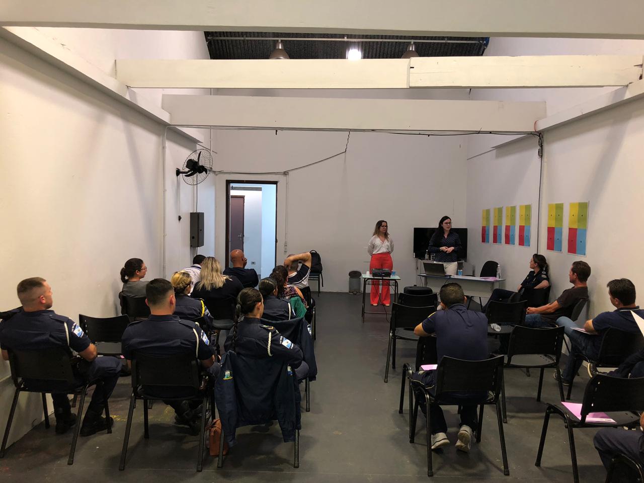 Na imagem vemos a equipe da DPPR palestrando aos guardas municipais. A equipe está de pé, ao fundo da sala, na frente dos oficias (esses estão sentados de costas).