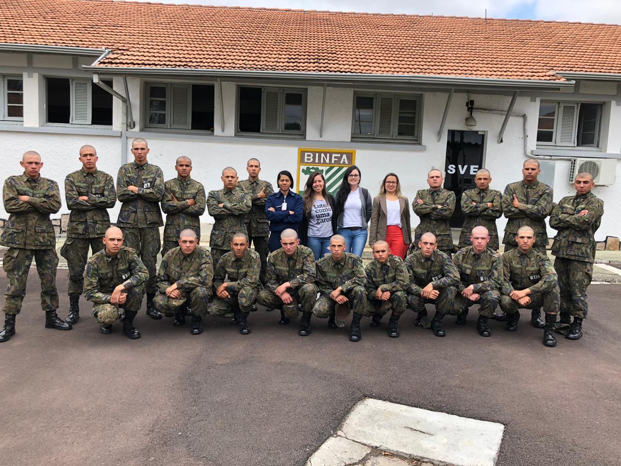 Vemos os soldados, alunos do curso, em fileiras - alguns abaixados e outros de pé - posando para foto. A equipe da DPPR da Casa da Mulher Brasileira se encontra em pé, no meio dos soldados,