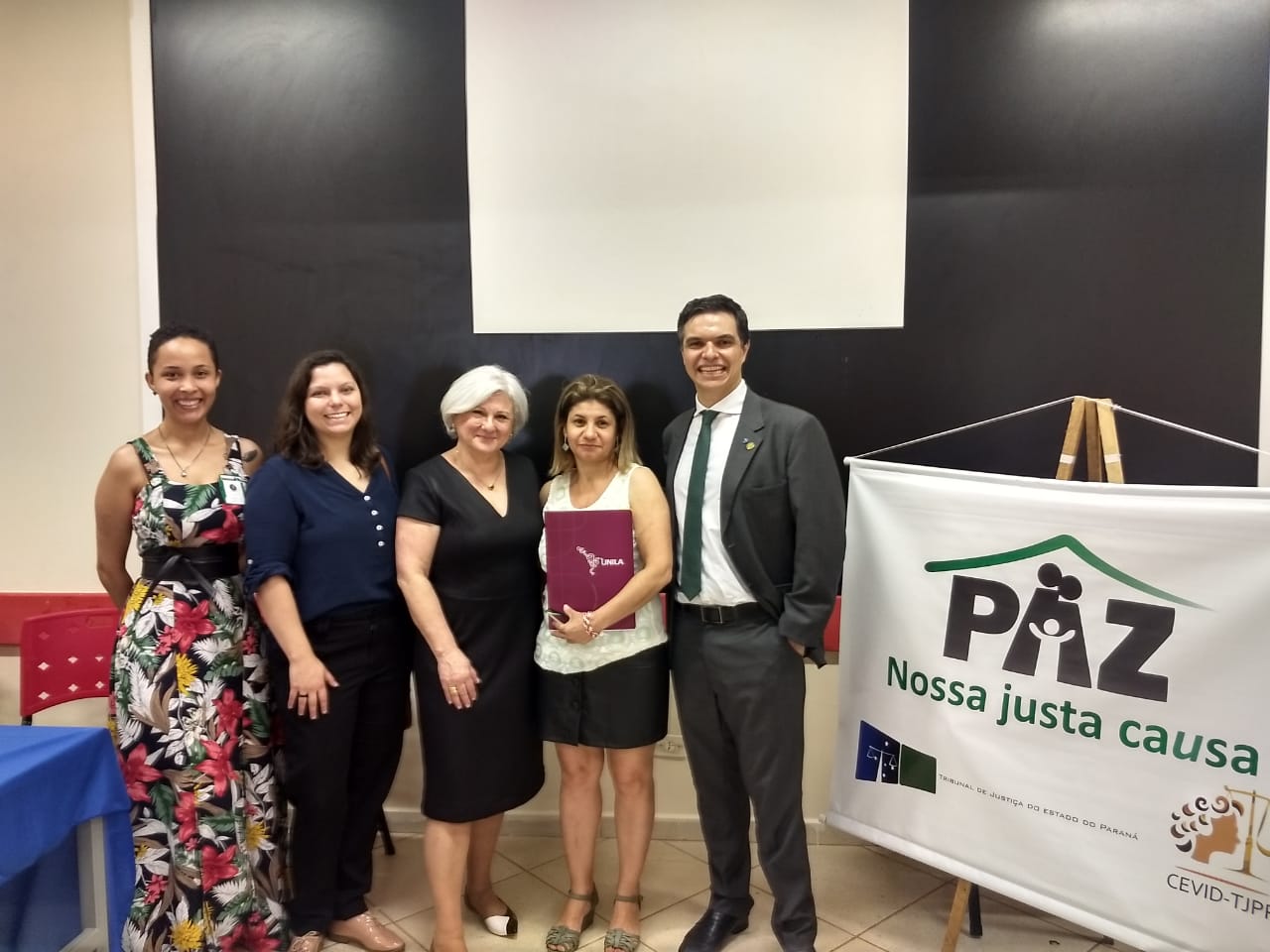 Na imagem vemos a Equipe Técnica da DPPR juntos, posando para a foto, do lado de um cartaz do evento.