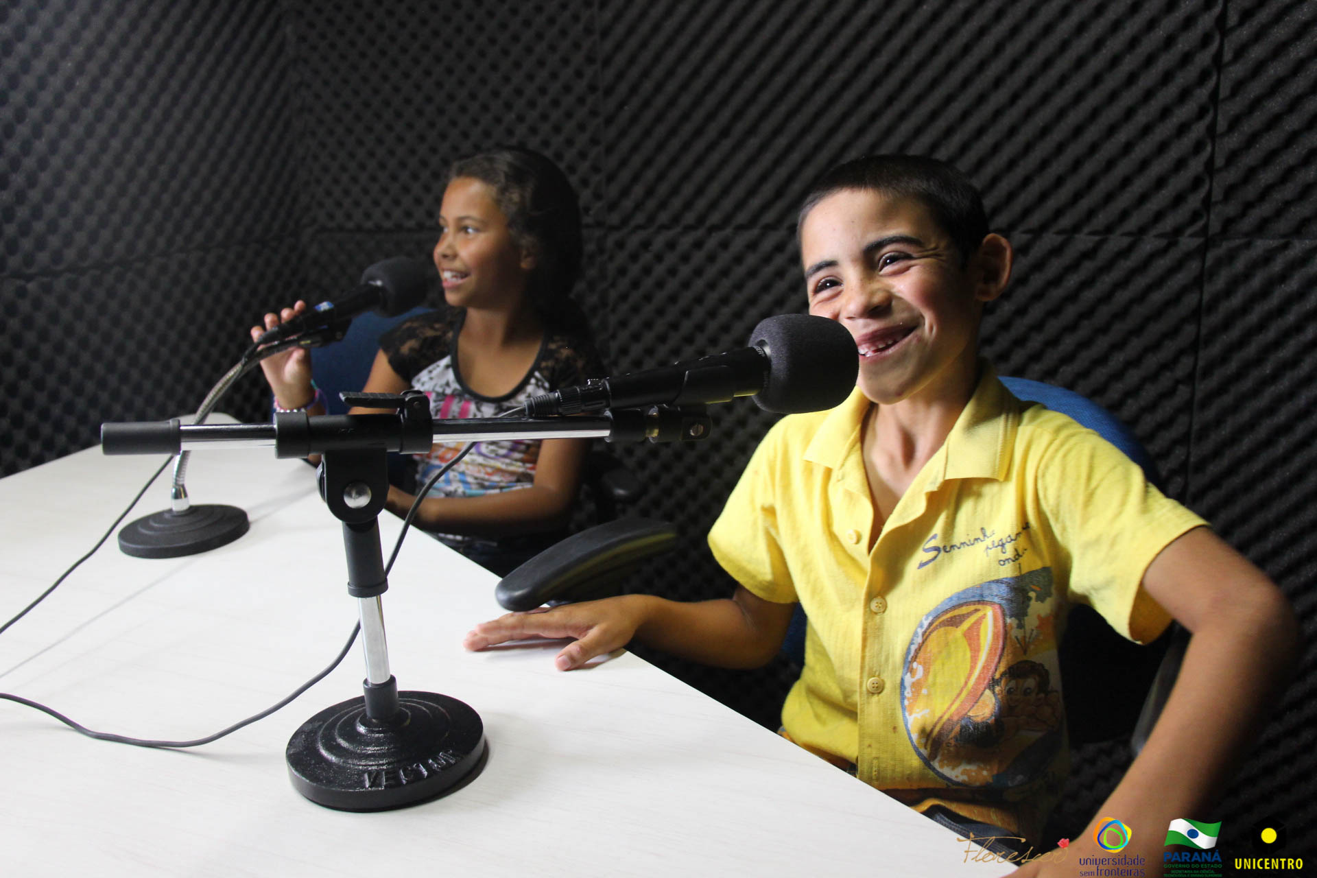Na imagem vemos duas crianças em um estúdio de rádio, sentadas na mesa de gravação.