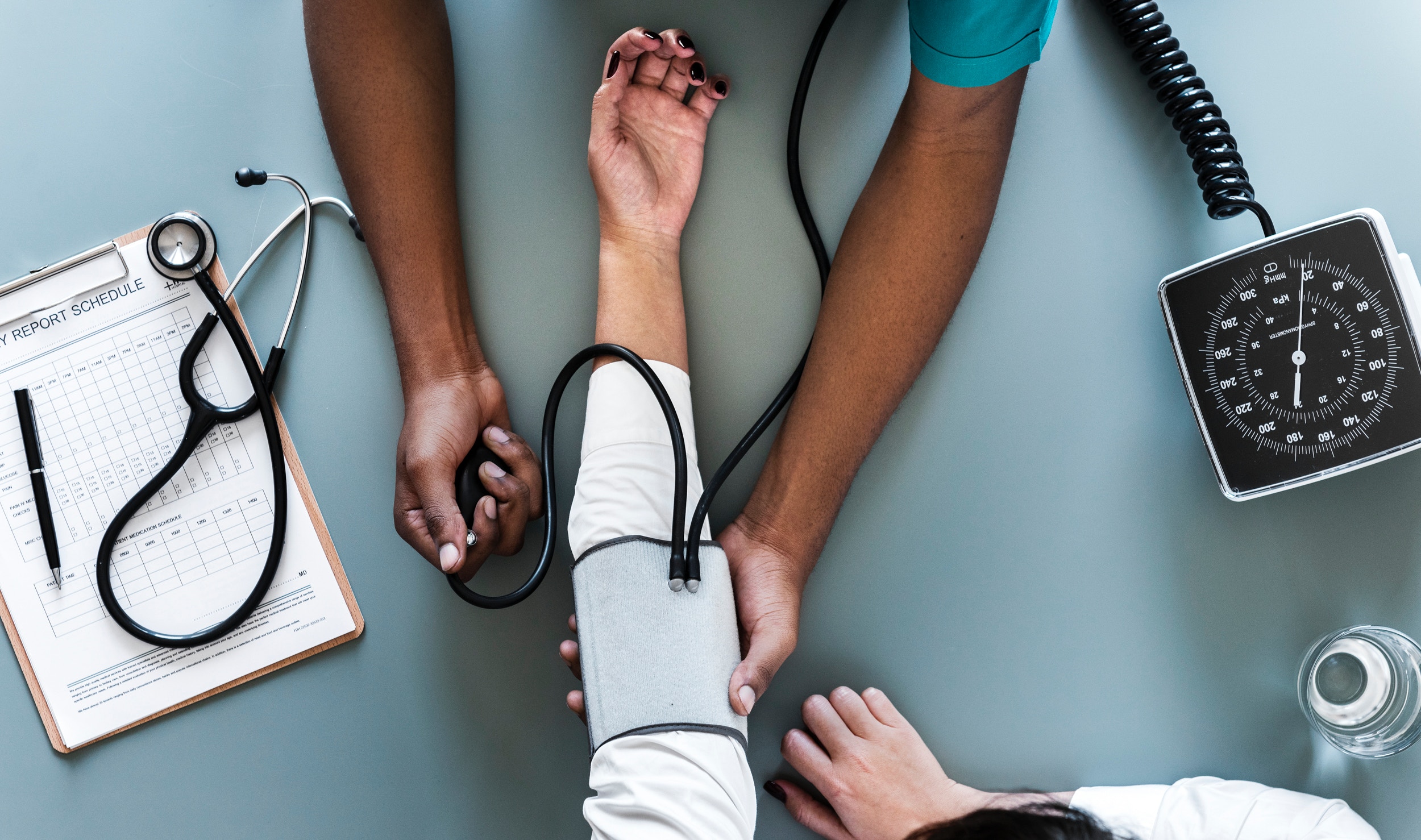 Imagem de um médico, onde esse está medindo a pressão do paciente.
A foto encontra-se em uma perceptiva observada de cima, aparecendo apenas os braços de ambos.