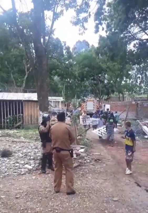 Imagem da reintegração de posse. Policiais no canto esquerdo da foto, no direito estão as pessoas desabrigadas carregando suas coisas.