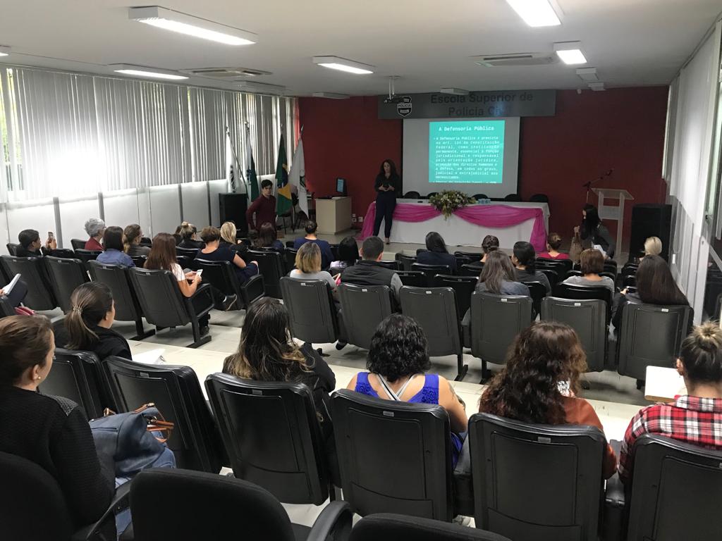 Imagem tirada durante a palestra do funda da sala. Vemos as costas dos ouvintes e a palestrante a frente.