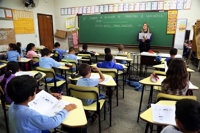 #LegendaAcessível: A imagem mostra a professora, na frente do quadro-negro ensinando para uma sala cheia de alunos, que acompanham a matéria pela apostila.