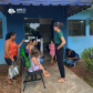 Foto de uma servidora branca com calça preta e camiseta da instituição na cor verde, conversando com usuárias da defensoria que esperam sentadas em cadeiras do lado de fora do prédio do CRAS. O prédio é de cor azul, em frente estão três cadeiras verdes e uma preta, onde estão sentadas duas crianças acompanhadas de suas mães, sendo elas uma mulher negra e duas mulheres brancas. Mais ao lado, uma criança branca aguarda ao lado da porta e, agachado também ao lado da porta, um homem mexe no celular.