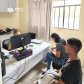 Foto de uma servidora amarela com a camiseta da instituição mexendo em um computador enquanto atende dois usuários, sendo eles um homem negro de camisa preta e branca e calça jeans, e uma mulher branca com calça jeans e camiseta cinza mexendo no celular.