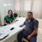 Imagem de uma servidora da defensoria com pele branca e camiseta da instituição na cor verde, com os braços apoiados em uma mesa branca. Ela atende um usuário branco com camiseta e calça em tons de azul. Ele segura uma caneta para assinar alguns papeis que estão sobre a mesa.