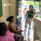 Imagem de duas mulheres negras usuárias da defensoria sentadas na parte interna do prédio do CRAS sendo atendidas por uma servidora branca que usa a camiseta da instituição e calça jeans.