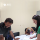 Imagem de uma servidora da Defensoria com a camisa da instituição em verde entregando documento para um homem branco e, ao fundo, uma mullher branca assina alguns papeis em uma mesa branca.