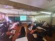 Imagem de uma das oficinas do evento que aconteceu em uma sala de aula. As pessoas formaram um círculo com as carteiras e estão atentas ao que fala uma das participantes.