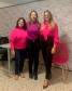 Na foto estão pousando as secretárias de atendimento Daiane Cristiane Pereira dos Santos e Dioneia dos Santos Modesto, e a defensora pública de Paranaguá Ingrid Lima Vieira, nesta ordem da esquerda para a direitas. As três estão sorrindo, com roupas cor de rosa e roxa. 