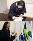 Imagem em formato de colagem, na imagem acima, uma foto retrato da nova Defensora Pública assinando seu termo de exercício como Defensora Pública, na imagem abaixo, mostra ela discursando ao lado de três bandeiras, sendo a primeira uma bandeira do Paraná, a segunda, uma bandeira do Brasil e a terceira sendo a bandeira da Defensoria Pública do Estado do Paraná.