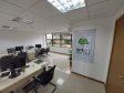 Imagem que mostra outra sala da DPE-PR no Fórum de Cambé, onde há um banner com o logo da Defensoria na parede ao lado da porta e três mesas com computadores e cadeiras para pessoas serem atendidas. Ao fundo é possível ver uma grande janela que mostra uma parte externa do Fórum, onde há algumas plantas em um canteiro.