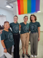 Imagem de uma foto espontânea de servidoras públicas, todas usando uma camiseta verde da Defensória Pública do Estado do Paraná. No fundo da imagem, dá para ver uma bandeira LGBTQIA+ pendura do teto.