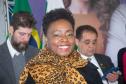 Na imagem se vê uma mulher negra sorrindo em primeiro plano e dois homens desfocados ao fundo. 
