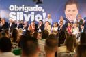 Imagem colorida horizontal. No primeiro plano se vê o público observando autoridades no lançamento do projeto de construção do Centro de Reabilitação Silvio Santos. Ao fundo cartaz com a imagem do apresentador onde se lê: Obrigado, Silvio.
