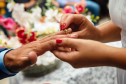 Imagem que mostra uma das noivas colocando a aliança no dedo do noivo. O fundo mostra flores desfocadas. É possível ver parte do vestido dela, branco. Ela tem as unhas pintadas com esmalte vermelho.
